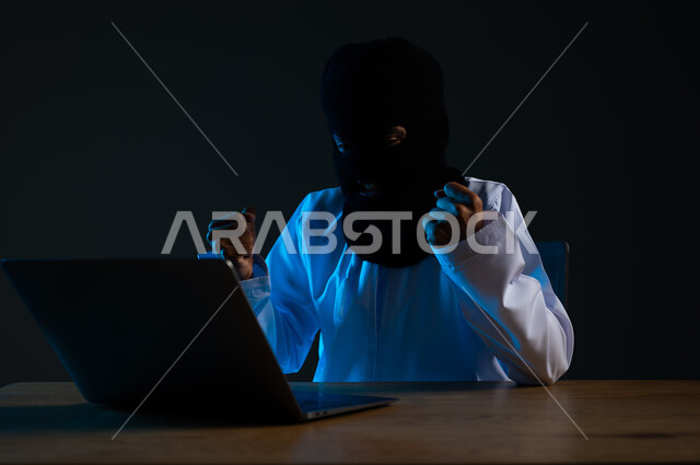 القدرة على تهكير الأجهزة, استخدم جهاز الحاسوب لأغراض غير مشروعة, مفهوم الجرائم الالكترونية, قرصنة وسرقة البيانات, تدمير الحسابات الرقمية, بورتريه مقرب لهاكر عربي خليجي عماني ملثم يرتدي الدشداشة يرفع قبضة يديه بإيماءات الفرح لنجاح عملية الاختراق، خلفية سوداء