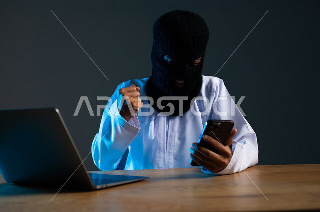 التجسس على المحادثات والحسابات الشخصية, سرقة وتهكير البيانات الالكترونية, استخدام الحاسوب المحمول لأغراض غير قانونية, بورتريه مقرب لهاكر عربي خليجي عماني ملثم يرتدي الدشداشة يرفع قبضة يده بإيماءات الفرح لنجاح عملية الاختراق، خلفية رمادية