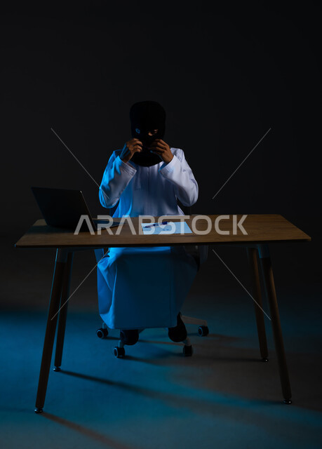 مفهوم الجرائم الالكترونية, القدرة على تهكير الأجهزة, استخدم جهاز الحاسوب لأغراض غير مشروعة, قرصنة وسرقة البيانات, تدمير الحسابات الرقمية, بورتريه لهاكر عربي خليجي عماني ملثم يرتدي الدشداشة يرفع يديه بتعابير الفرح، خلفية سوداء