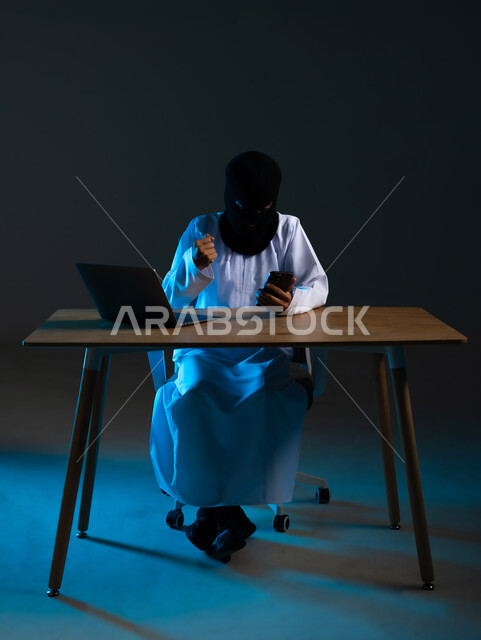 قرصنة وسرقة البيانات, التسلل عبر الأمن السيبراني, خوارزميات لتدمير الحسابات الرقمية, استخدم جهاز الحاسوب المحمول لأغراض غير قانونية, بورتريه لهاكر عربي خليجي عماني ملثم يرتدي الدشداشة يرفع قبضة يده بإيماءات الفرح لنجاح عملية الاختراق, خلفية زرقاء