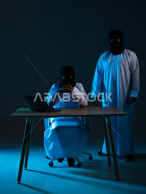 استخدم جهاز الحاسوب لأغراض غير قانونية, التسلل عبر الأمن السيبراني، قرصنة وسرقة البيانات, خوارزميات لتدمير الحسابات الرقمية, بورتريه لهكرين عربيين خليجيين عمانيين ملثمين يرتديان الدشداشة يعملان في مجال الهكر, خلفية زرقاء