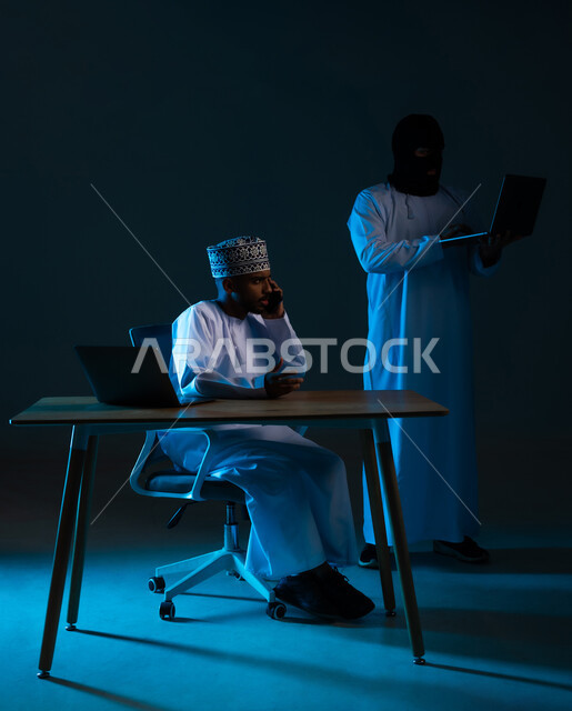 Spying on phone calls, stealing and hacking electronic data, hacking systems and protecting devices, exposure to attacks and hacking, portrait of a hacker using a computer to hack the phone of an Arab Gulf Omani man wearing a dishdasha and a kummah, blue background