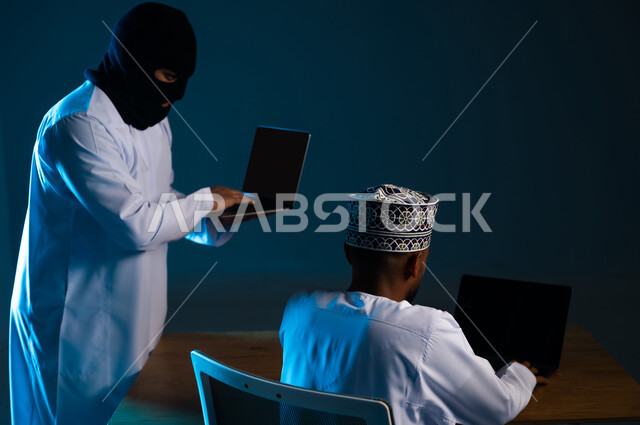 Hacking user protection and security, exposure to attacks and hacks, destruction of digital accounts, infiltration through cyber security, portrait of a hacker using a laptop to hack the device of an Arab Gulf Omani man wearing a dishdasha and a kemah, displaying a blank black screen through the computer, blue background