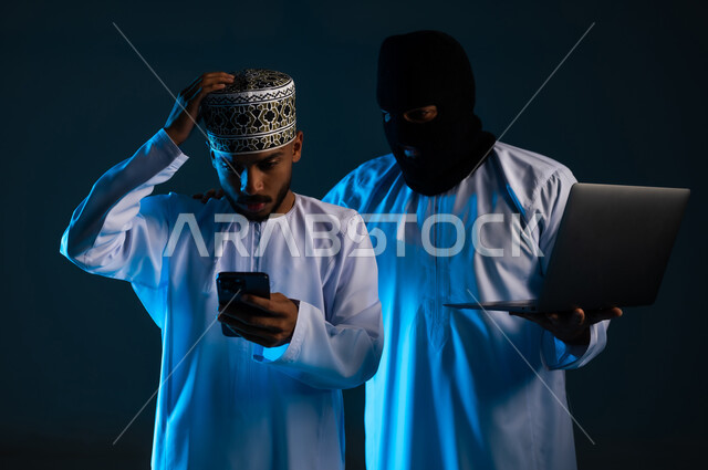 التجسس على المحادثات والحسابات الشخصية, سرقة وقرصنة البيانات الالكترونية, اختراق الأنظمة وحماية وأمن الأجهزة, التعرض للهجمات والاختراقات, بورتريه مقرب لهاكر يستخدم جهاز الحاسوب لاختراق هاتف رجل عربي خليجي عماني يرتدي الدشداشة والكمة، خلفية زرقاء