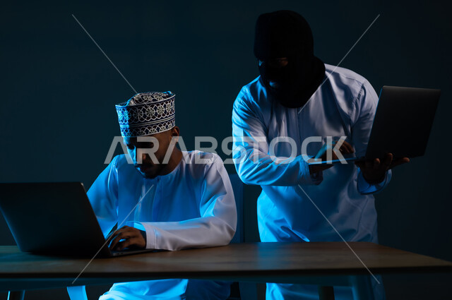 تدمير الحسابات الرقمية, التسلل عبر الأمن السيبراني، قرصنة وسرقة البيانات الشخصية, التعرض للهجمات والاختراقات، اختراق حماية وأمن وخصوصية المستخدمين, بورتريه لهاكر يستخدم الحاسوب المحمول لاختراق جهاز رجل عربي خليجي عماني يرتدي الدشداشة والكمة, خلفية زرقاء