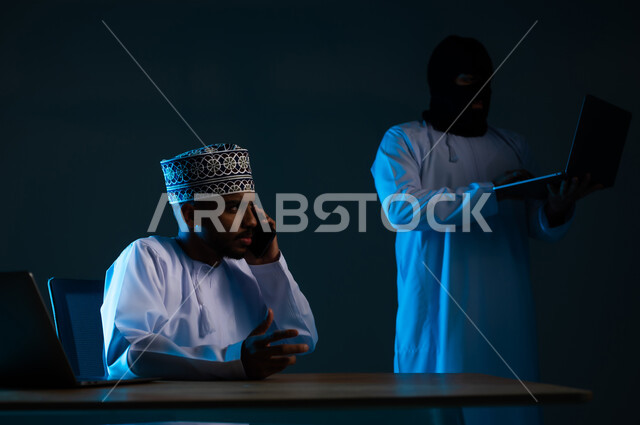 Spying on phone calls, stealing and hacking electronic data, hacking systems and protecting and securing devices, exposure to attacks and hacking, close-up portrait of a hacker using a computer to hack the phone of an Arab Gulf Omani man wearing a dishdasha and a kimmah, blue background