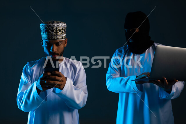 التجسس على المحادثات والحسابات الشخصية, سرقة وقرصنة البيانات, اختراق الأنظمة وحماية وأمن الأجهزة, التعرض للهجمات والاختراقات, بورتريه مقرب لهاكر يستخدم جهاز الحاسوب لاختراق هاتف رجل عربي خليجي عماني يرتدي الدشداشة والكمة، خلفية زرقاء