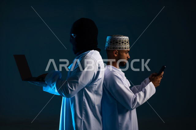 Spying on conversations and personal accounts, data theft and hacking, hacking systems and protecting and securing devices, exposure to attacks and hacking, close-up portrait from the side of a hacker using a computer to hack the phone of an Arab Gulf Omani man wearing a dishdasha and a kummah, blue background