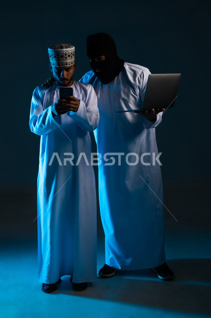 The concept of cybercrime, the ability to hack devices, exposure to attacks and breaches, breaching the protection, security and privacy of users, portrait of a hacker using a computer to hack the phone of an Arab Gulf Omani man wearing a dishdasha and a cloak, full-length body, blue background