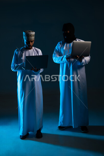 Exposure to attacks and hacks, hacking of users' protection, security and privacy, concept of cybercrime, ability to hack devices, portrait of a hacker using a laptop to hack the device of an Arab Gulf Omani man wearing a dishdasha and a kimono, full-length body, blue background