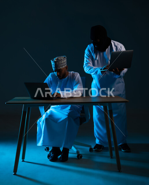 Data theft and hacking, system hacking and device protection and security, exposure to attacks and hacking, concept of cybercrime, portrait of a hacker using a laptop to hack the device of an Arab Gulf Omani man wearing a dishdasha and a kimono, blue background