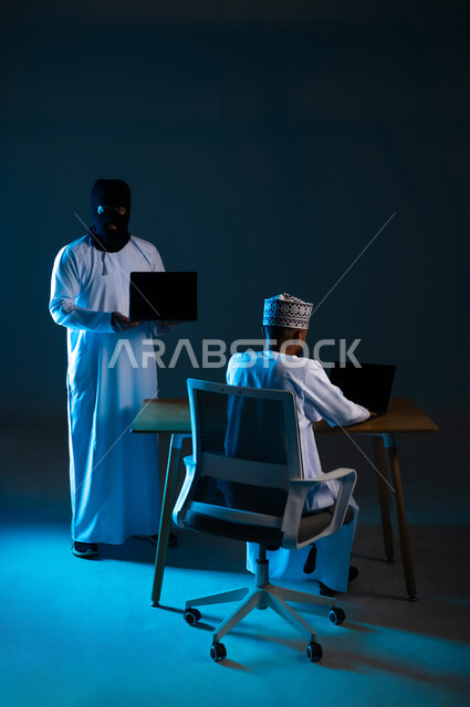 Hacking systems and protecting and securing devices, exposure to attacks and hacking, data theft and piracy, portrait of a hacker using a laptop to hack the device of an Arab Gulf Omani man wearing a dishdasha and a turban, displaying a blank black screen via computer, blue background