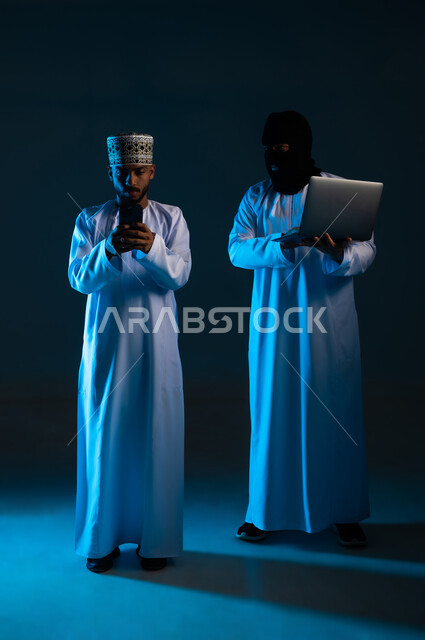 Ability to hack devices, exposure to attacks and breaches, breach of protection, security and privacy of users, concept of cybercrime, portrait of a hacker using a computer to hack the phone of an Arab Gulf Omani man wearing a dishdasha and a kimono, full body, blue background