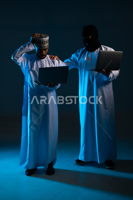 نجاح عملية الاختراق, التعرض للهجمات والاختراقات، مفهوم الجرائم الالكترونية, القدرة على تهكير الأجهزة, بورتريه لهاكر يستخدم الحاسوب المحمول لاختراق جهاز رجل عربي خليجي عماني يرتدي الدشداشة والكمة، الجسم بالطول الكامل, خلفية زرقاء