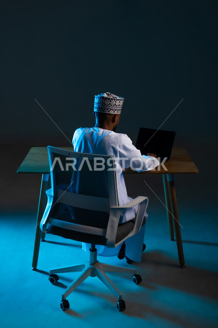 سرقة البيانات السرية، اختراق الأنظمة وحماية الأجهزة, التعرض للهجمات والاختراقات, مفهوم الجرائم الالكترونية, بورتريه من الخلف لرجل عربي خليجي عماني يرتدي الدشداشة والكمة يستعمل الحاسوب المحمول في الهكر الغير قانوني, عرض شاشة سوداء فارغة عبر الحاسب, خلفية زرقاء