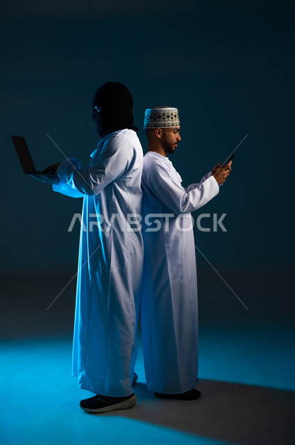 Concept of cybercrime, ability to hack devices, exposure to attacks and breaches, breach of user protection and security, side portrait of a hacker using a computer to hack the phone of an Arab Gulf Omani man wearing a dishdasha and a kimono, full-length body, blue background