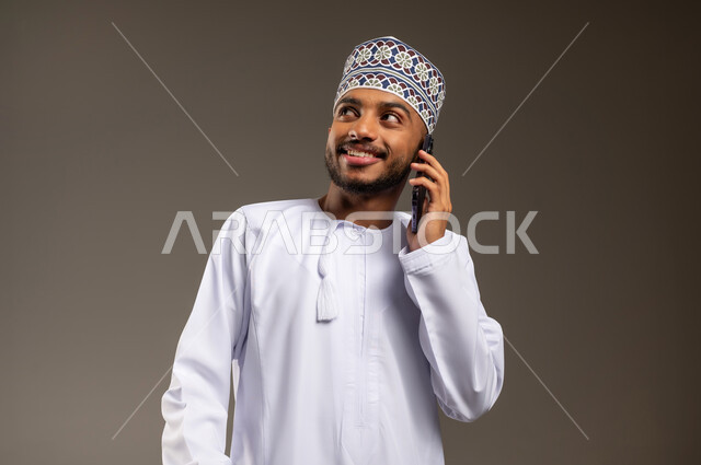 Using a modern and advanced technical device, communicating with family and friends, making a phone call via mobile, close-up portrait of a smiling young Arab Gulf Omani man wearing a dishdasha and a kummah looking at something above and holding a mobile phone in his hand, gray background
