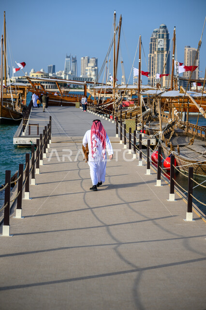 الاستمتاع بالنزهات البحرية الهادئة، مهرجان كتارا الرابع عشر للمحامل، الواجهة البحرية في دولة قطر في النهار، أماكن سياحية لقضاء أوقات ممتعة، صورة من الخلف لرجل عربي خليجي قطري يرتدي الثوب التقليدي والشماغ يمشي على كورنيش مدينة الدوحة