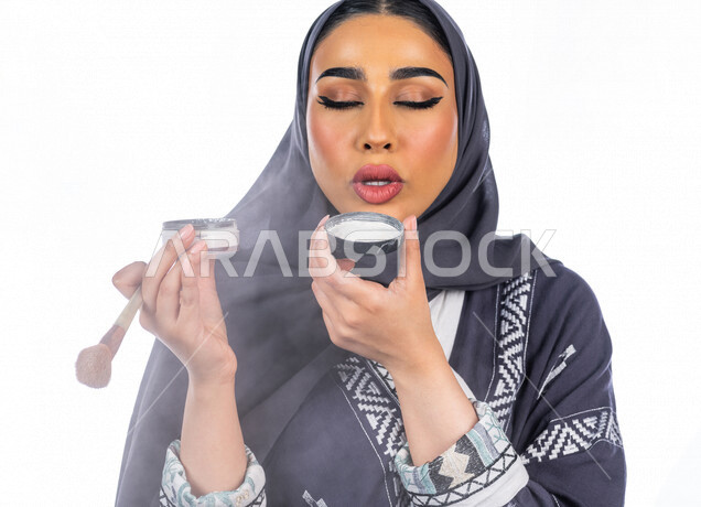 بورتريه لمرأة عربية خليجية سعودية تحمل إحدى أدوات التجميل (الفرشاة) على بشرة الوجه، خلفية بيضاء لمرأة خبيرة بمستحضرات التجميل، التسويق لمنتجات التجميل، البيع عبر الانترنت، متجر بيع مستحضرات التجميل، مفهوم الإهتمام بالجمال والبشرة، ادوات مكياج تتوريال