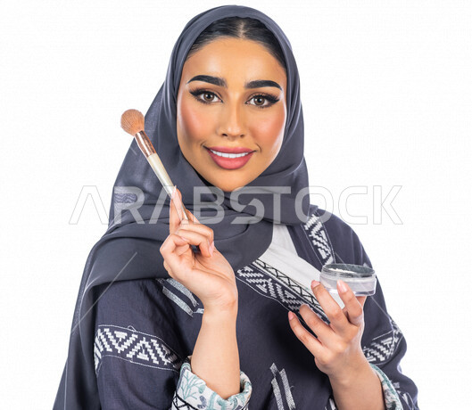 بورتريه لمرأة عربية خليجية سعودية تحمل إحدى أدوات التجميل (الفرشاة) على بشرة الوجه، خلفية بيضاء لمرأة خبيرة بمستحضرات التجميل، التسويق لمنتجات التجميل، البيع عبر الانترنت، متجر بيع مستحضرات التجميل، مفهوم الإهتمام بالجمال والبشرة، ادوات مكياج تتوريال