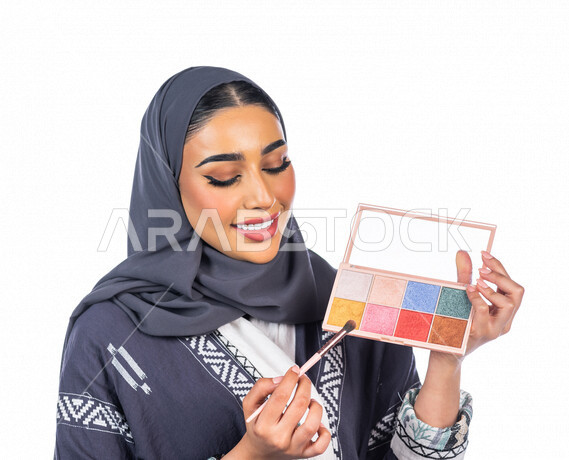 بورتريه لمرأة عربية خليجية سعودية تحمل إحدى أدوات التجميل (فرشاة ظل العيون) على بشرة الوجه، خلفية بيضاء لمرأة خبيرة بمستحضرات التجميل، التسويق لمنتجات التجميل، متجر بيع مستحضرات التجميل، مفهوم الإهتمام بالجمال والبشرة، ادوات مكياج تتوريال