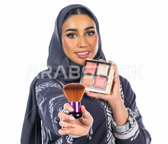 بورتريه لمرأة عربية خليجية سعودية تحمل إحدى أدوات التجميل (الفرشاة) على بشرة الوجه، خلفية بيضاء لمرأة خبيرة بمستحضرات التجميل، التسويق لمنتجات التجميل، البيع عبر الانترنت، متجر بيع مستحضرات التجميل، مفهوم الإهتمام بالجمال والبشرة، ادوات مكياج تتوريال