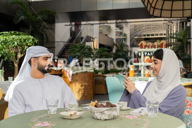 استغلال وقت الفراغ بالتسلية، اجواء اسرية سعيدة مليئة بالحب، مفهوم الألفة والمحبة بين أفراد الأسرة, صورة مقربة لرجل عربي خليجي اماراتي يرتدي الكندورة والغترة يجلس باحدى الكافيهات تقوم زوجته بالتقاط صور تذكاريه