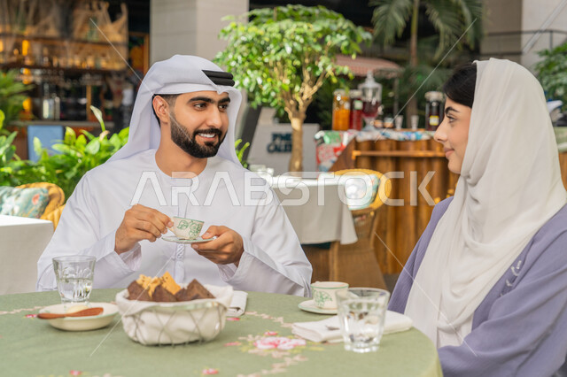 الاستمتاع بتناول المشروبات الساخنة، حوارات ونقاشات عائلية، قضاء وقت ممتع في الخارج، مفهوم الألفة والمحبة بين أفراد الأسرة, صورة مقربة لرجل عربي خليجي اماراتي يرتدي الكندورة والغترة يجلس برفقة زوجته في احدى الكافيهات يقوم بشرب القهوة