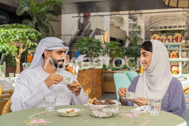 الاستمتاع بتناول المشروبات الساخنة، حوارات ونقاشات عائلية، قضاء وقت ممتع في الخارج، مفهوم الألفة والمحبة بين أفراد الأسرة, صورة مقربة لرجل عربي خليجي اماراتي يرتدي الكندورة والغترة يجلس برفقة زوجته في احدى الكافيهات يقوم بشرب القهوة