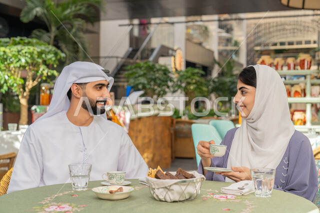 الاستمتاع بتناول المشروبات الساخنة، حوارات ونقاشات عائلية، قضاء وقت ممتع في الخارج، مفهوم الألفة والمحبة بين أفراد الأسرة, صورة مقربة لرجل عربي خليجي اماراتي يرتدي الكندورة والغترة يجلس برفقة زوجته في احدى الكافيهات يقوم بشرب القهوة