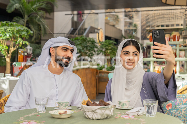 أجواء اسرية سعيدة مليئة بالحب، قضاء وقت ممتع في الخارج، استخدام الاجهزة التقنية الحديثة، مفهوم الألفة والمحبة بين أفراد الأسرة, صورة مقربة لرجل عربي خليجي اماراتي يرتدي الكندورة والغترة يجلس باحدى الكافيهات يقوم بالتقاط صور سيلفي تذكارية