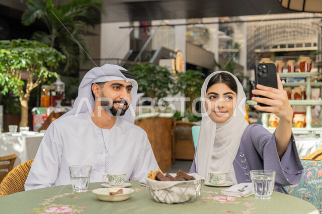 أجواء اسرية سعيدة مليئة بالحب، قضاء وقت ممتع في الخارج، استخدام الاجهزة التقنية الحديثة، مفهوم الألفة والمحبة بين أفراد الأسرة, صورة مقربة لرجل عربي خليجي اماراتي يرتدي الكندورة والغترة يجلس باحدى الكافيهات يقوم بالتقاط صور سيلفي تذكارية