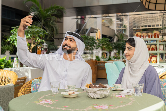أجواء اسرية سعيدة مليئة بالحب، قضاء وقت ممتع في الخارج، استخدام الاجهزة التقنية الحديثة، مفهوم الألفة والمحبة بين أفراد الأسرة, صورة مقربة لرجل عربي خليجي اماراتي يرتدي الكندورة والغترة يجلس باحدى الكافيهات يقوم بالتقاط صور سيلفي تذكارية
