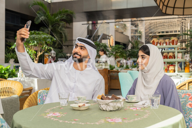 أجواء اسرية سعيدة مليئة بالحب، قضاء وقت ممتع في الخارج، استخدام الاجهزة التقنية الحديثة، مفهوم الألفة والمحبة بين أفراد الأسرة, صورة مقربة لرجل عربي خليجي اماراتي يرتدي الكندورة والغترة يجلس باحدى الكافيهات يقوم بالتقاط صور سيلفي تذكارية