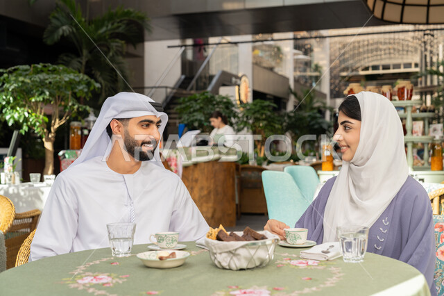 الاستمتاع بتناول المشروبات الساخنة، حوارات ونقاشات عائلية، قضاء وقت ممتع في الخارج، مفهوم الألفة والمحبة بين أفراد الأسرة, صورة مقربة لرجل عربي خليجي اماراتي يرتدي الكندورة والغترة يجلس برفقة زوجته في احدى الكافيهات يقوم بشرب القهوة