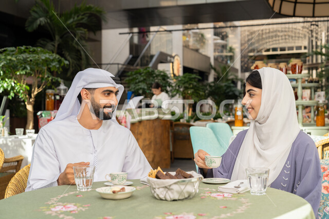 الاستمتاع بتناول المشروبات الساخنة، حوارات ونقاشات عائلية، قضاء وقت ممتع في الخارج، مفهوم الألفة والمحبة بين أفراد الأسرة, صورة مقربة لرجل عربي خليجي اماراتي يرتدي الكندورة والغترة يجلس برفقة زوجته في احدى الكافيهات يقوم بشرب القهوة