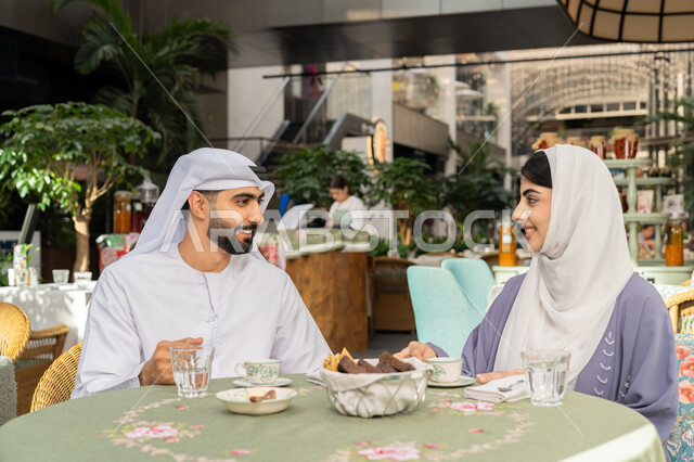 الاستمتاع بتناول المشروبات الساخنة، حوارات ونقاشات عائلية، قضاء وقت ممتع في الخارج، مفهوم الألفة والمحبة بين أفراد الأسرة, صورة مقربة لرجل عربي خليجي اماراتي يرتدي الكندورة والغترة يجلس برفقة زوجته في احدى الكافيهات يقوم بشرب القهوة
