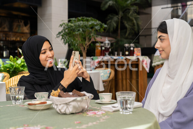 قضاء أوقات ممتعة مع الأصدقاء،  تناول المشروبات الساخنة والحلويات في الامارات، صورة مقربة لإمرأة عربية خليجية اماراتية ترتدي الحجاب والعباءة السوداء تقوم بالتقاط صورة تذكارية لصديقتها عبر الهاتف المحمول،  شابة سعودية تقوم بتوثيق اللحظات المميزة مع صديقتها