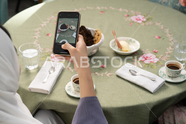 Having a good time in a restaurant, using a modern and advanced technical device, a close-up photo from the back of a veiled Emirati Gulf Arab woman wearing an abaya sitting on a comfortable chair, a young Saudi woman taking a souvenir photo via mobile, eating hot drinks and sweets in restaurants and cafes in the Emirates