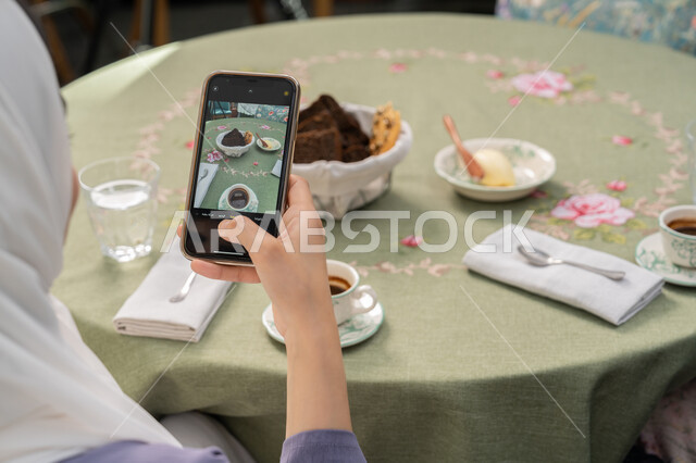 Using a modern and advanced technical device, having a good time in a restaurant, a close-up photo from the back of a veiled Emirati Gulf Arab woman wearing an abaya sitting on a comfortable chair, a young Saudi woman taking a souvenir photo via mobile, eating hot drinks and sweets in restaurants and cafes in the Emirates