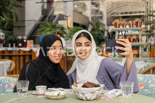 Documenting happy moments by taking a souvenir selfie, a close-up photo of an Emirati Gulf Arab woman wearing a hijab and abaya sitting on the couch with her friend in a cafe and using a mobile phone, sharing hot drinks and sweets, two young Saudi women having a fun and entertaining time