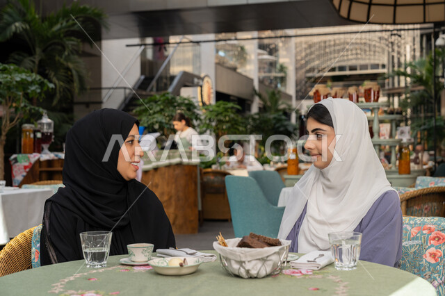 النقاشات والحوارات في الأجواء الهادئة، بناء علاقات اجتماعية ناجحة، شابتان سعوديتان تقضي اوقات ممتعة، صورة مقربة لإمرأة عربية خليجية اماراتية ترتدي الحجاب والعباءة السوداء تجلس على الأريكة مع صديقتها بإحدى المقاهي بإيماءات السعادة، مشاركة المشروبات الساخنة