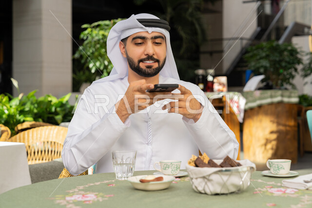 توثيق اللحظات السعيدة، قضاء أوقات ممتعة في المطعم، رجل عربي خليجي اماراتي يرتدي الكندورة والغترة البيضاء يجلس على مائدة الطعام يمسك بيده الهاتف المحمول يقوم بالتقاط الصور للسفرة، مطاعم و كافيهات في الإمارات العربية المتحدة