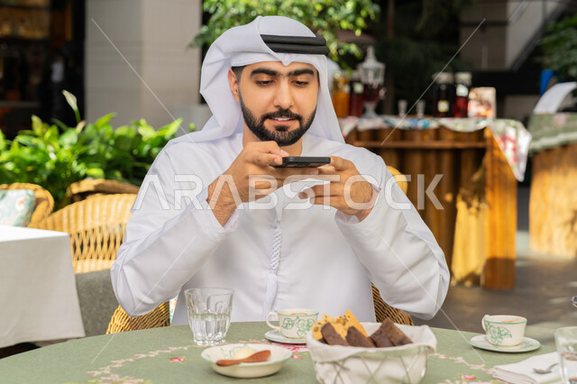قضاء أوقات ممتعة في المطعم، رجل عربي خليجي اماراتي يرتدي الكندورة والغترة البيضاء يجلس على مائدة الطعام يمسك بيده الهاتف المحمول يقوم بالتقاط الصور للسفرة، تشكيلة متنوعة من الحلويات والمشروبات الساخنة على الطاولة، مطاعم و كافيهات في الإمارات العربية المتحدة
