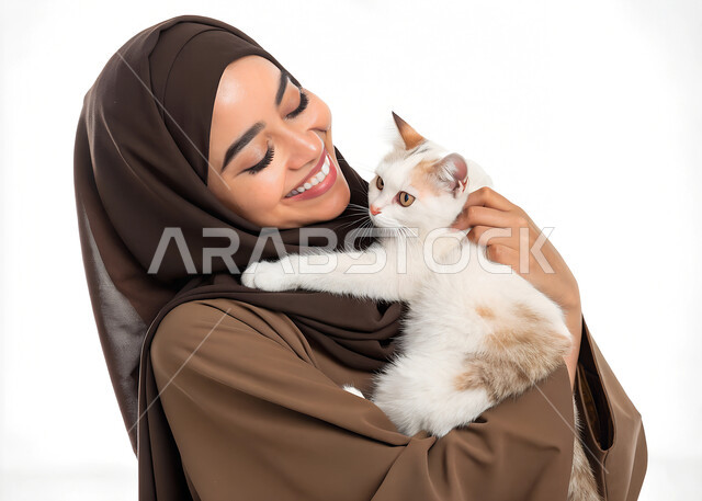 مفهوم الرفق بالحيوان والعطف عليه، الأثر الايجابي لاقتناء الحيوانات المنزلية وتربيتها، الشعور بالسعادة أثناء قضاء وقت ممتع في اللعب مع القط، بورتريه مقرب لإمرأة عربية خليجية سعودية مبتسمة ترتدي الحجاب والعباءة تقوم بحمل قطة أليفة، خلفية بيضاء