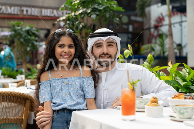 الاستعداد لالتقاط صورة تذكارية، فعاليات ترفيهية عائلية، الاحتفاظ بالذكريات السعيدة، مفهوم الألفة والمحبة بين أفراد الأسرة, قضاء وقت ممتع في الخارج، صورة مقربة لرجل عربي خليجي اماراتي يرتدي الكندورة والغترة يجلس في المقهى يقوم باحتضان ابنته