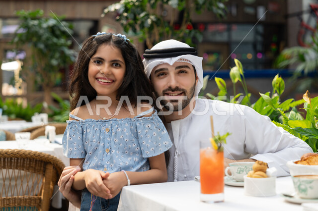 الاحتفاظ بالذكريات السعيدة، الاستعداد لالتقاط صورة تذكارية، فعاليات ترفيهية عائلية، مفهوم الألفة والمحبة بين أفراد الأسرة, قضاء وقت ممتع في الخارج، صورة مقربة لرجل عربي خليجي اماراتي يرتدي الكندورة والغترة يجلس في المقهى يقوم باحتضان ابنته
