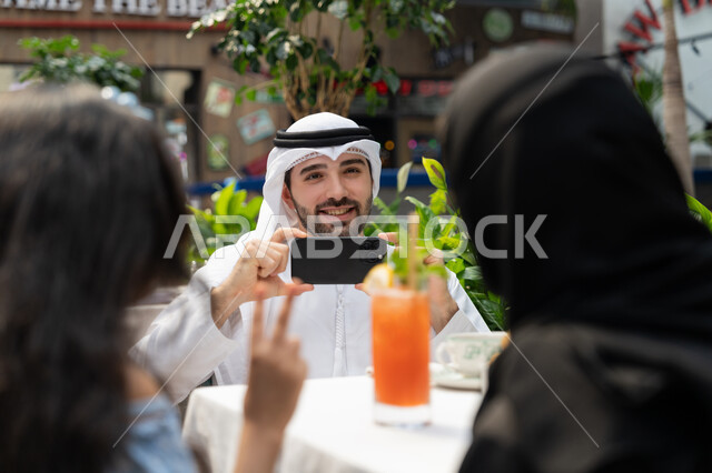 قضاء وقت ممتع في الخارج، فعاليات ترفيهية عائلية، الاحتفاظ بالذكريات السعيدة، مفهوم الألفة والمحبة بين أفراد الأسرة, صورة مقربة لرجل عربي خليجي اماراتي يرتدي الكندورة والغترة يجلس في المقهى يقوم بالتقاط صورة تذكارية لزوجته وابنته باستخدام الهاتف المحمول