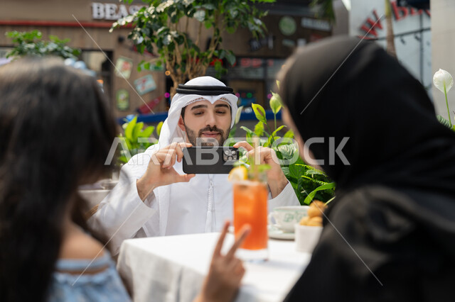 فعاليات ترفيهية عائلية، الاحتفاظ بالذكريات السعيدة، مفهوم الألفة والمحبة بين أفراد الأسرة, قضاء وقت ممتع في الخارج، صورة مقربة لرجل عربي خليجي اماراتي يرتدي الكندورة والغترة يجلس في المقهى يقوم بالتقاط صورة تذكارية لزوجته وابنته باستخدام الهاتف المحمول
