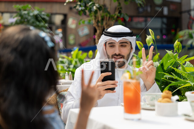 مفهوم الألفة والمحبة بين أفراد الأسرة، الاحتفاظ بالذكريات السعيدة، قضاء وقت ممتع في الخارج، صورة مقربة لرجل عربي خليجي اماراتي يرتدي الكندورة والغترة يجلس باحدى الكافيهات يرفع يده بايماءات النصر يقوم بالتقاط صورة تذكارية لابنته باستخدام الهاتف المحمول